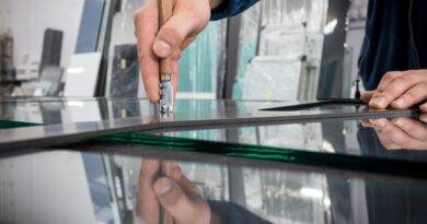 Ein Fensterbauer in Wuppertal schneidet präzise Glas mit einem speziellen Werkzeug. Der Handwerker trägt eine blaue Arbeitsjacke und arbeitet sorgfältig an einer Glasplatte in einer Werkstatt. Im Hintergrund sind weitere Glasplatten und Werkstattutensilien zu sehen.