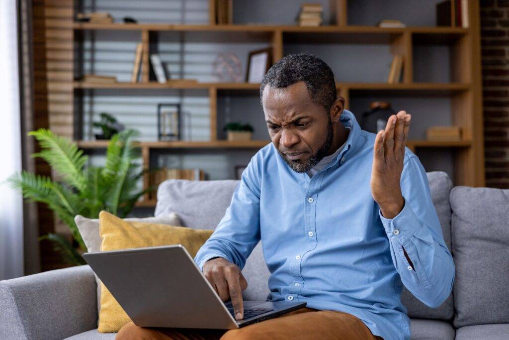 Frustrierter afroamerikanischer Mann mit Laptop, der technische Schwierigkeiten zu Hause hat