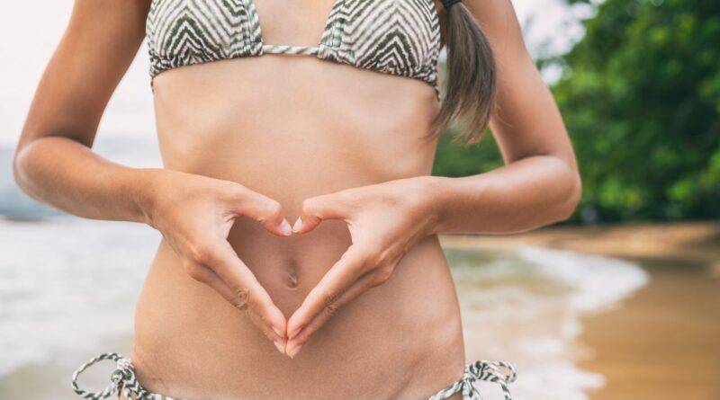 rau in einem Bikini am Strand formt ein Herz mit ihren Händen über ihrem Bauch, symbolisch für Körperbewusstsein und Selbstliebe nach einer erfolgreichen Kryolipolyse Behandlung