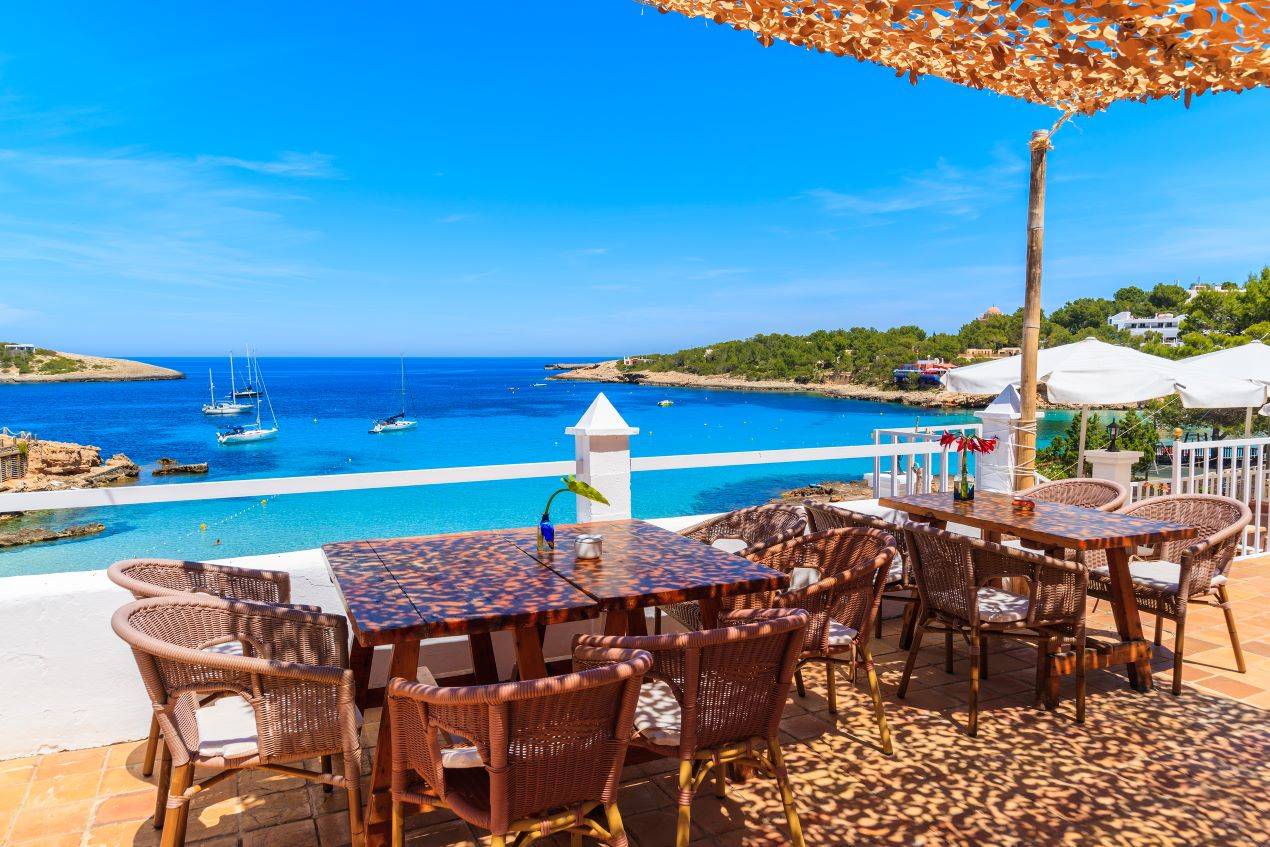 Gemütliche Terrasse eines Beach Restaurants auf Ibiza mit Blick auf das türkisfarbene Meer, Segelboote und eine idyllische Küstenlandschaft.