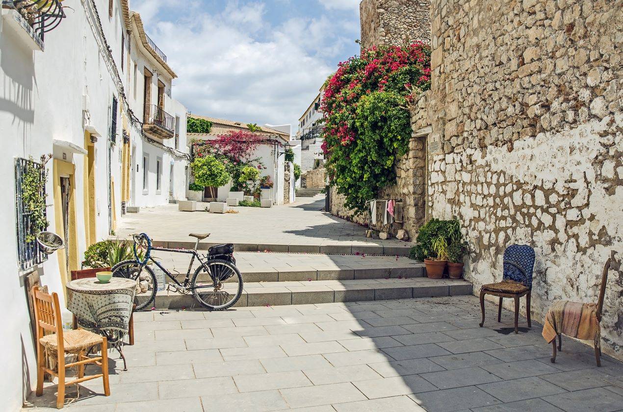 Pittoreske Gasse in der Altstadt von Ibiza mit Blumen geschmückten Häusern, einem Fahrrad und gemütlichen Sitzgelegenheiten – ideal als Ausflug vor dem Besuch eines Beach Restaurants Ibiza.