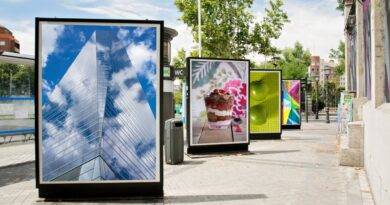 Werbedisplays auf einer städtischen Straße mit verschiedenen Bildern, darunter ein Hochhaus, Dessert, grüne Äpfel und bunte Muster, umgeben von Bäumen und urbaner Architektur.