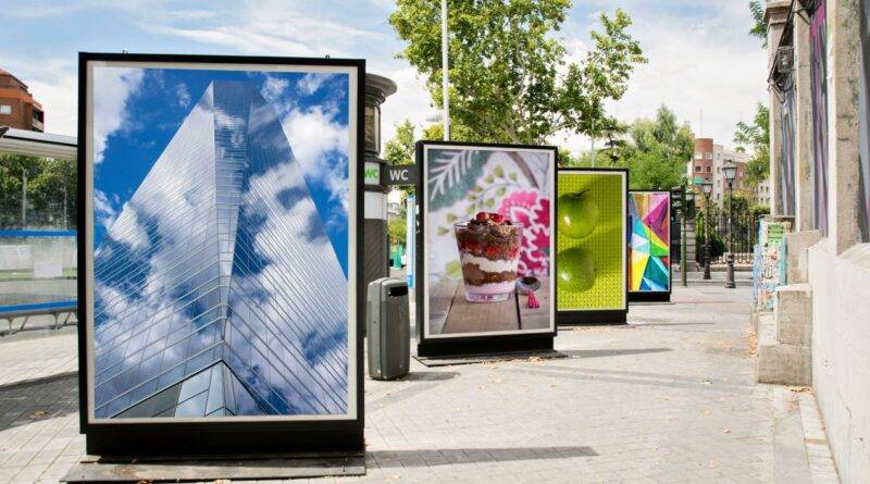 Werbedisplays auf einer städtischen Straße mit verschiedenen Bildern, darunter ein Hochhaus, Dessert, grüne Äpfel und bunte Muster, umgeben von Bäumen und urbaner Architektur.