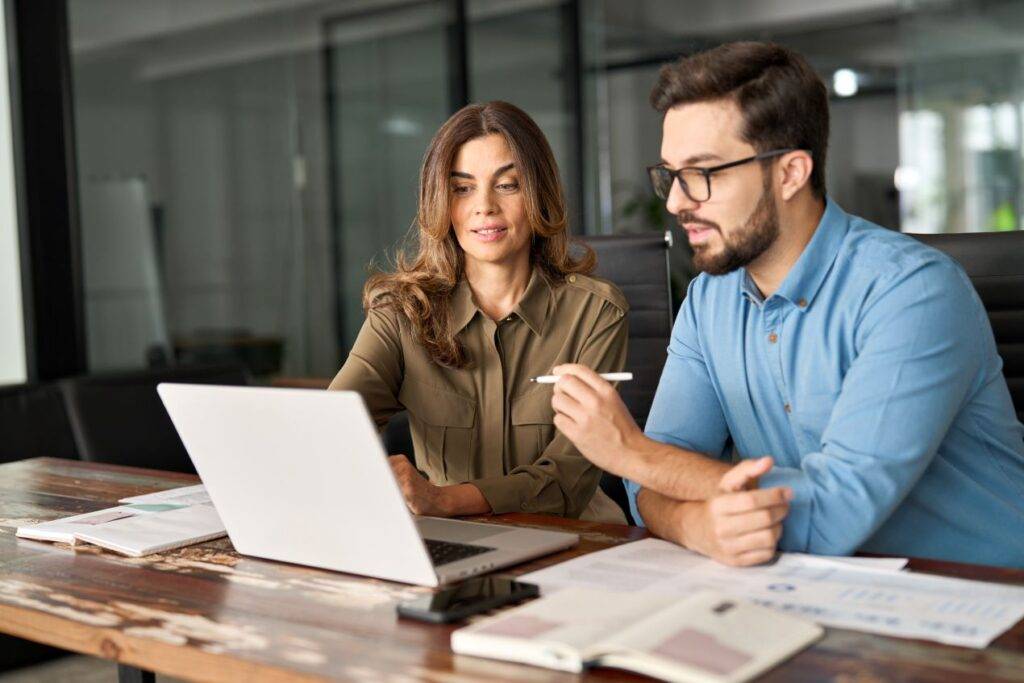 Zwei Geschäftspartner, eine Frau und ein Mann, arbeiten gemeinsam an einem Laptop in einem modernen Büro, während sie Event-Details und Optionen zur Display-Miete besprechen.